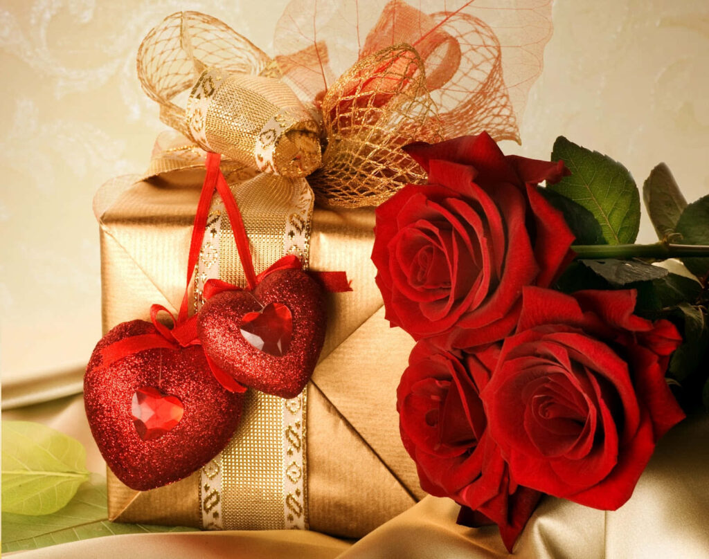 Gold gift box with decorative ribbon next to red roses and heart ornaments romantic scene wallpaper