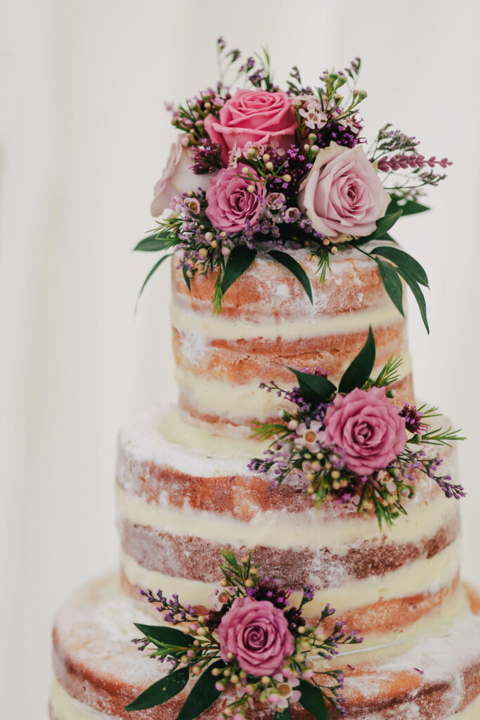 Multi-layered naked wedding cake with pink purple roses greenery and powdered sugar dusting wallpaper