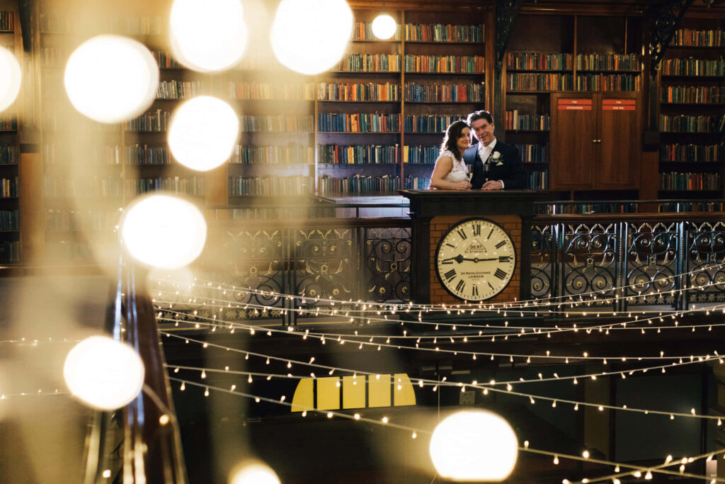 Newlywed couple embraces inside Adelaide State Library with warm string lights and bookshelves Wallpaper