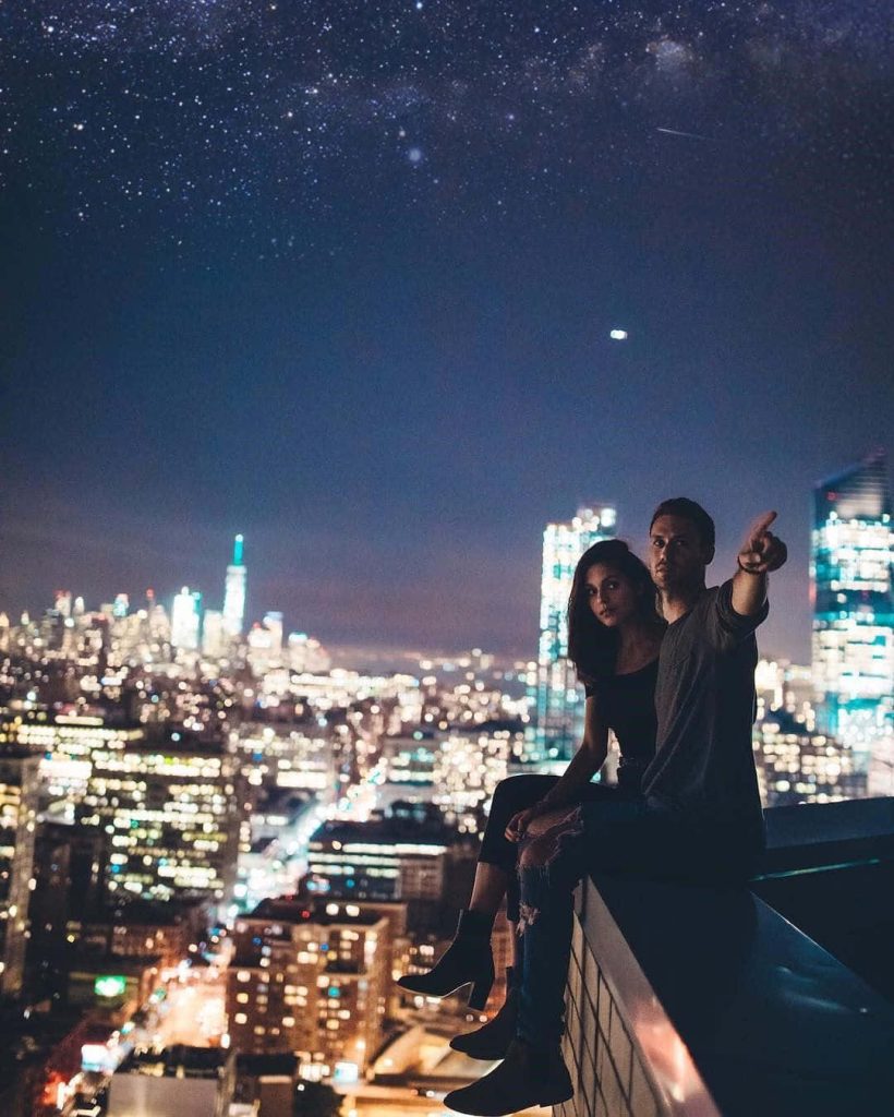 Young couple on rooftop ledge admiring star-lit city skyline at night romantic urban wallpaper