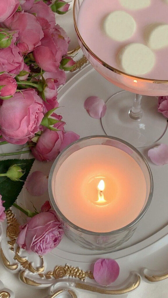 Rose pink candle glowing beside fresh pink roses petals and pastel drink on ornate tray wallpaper