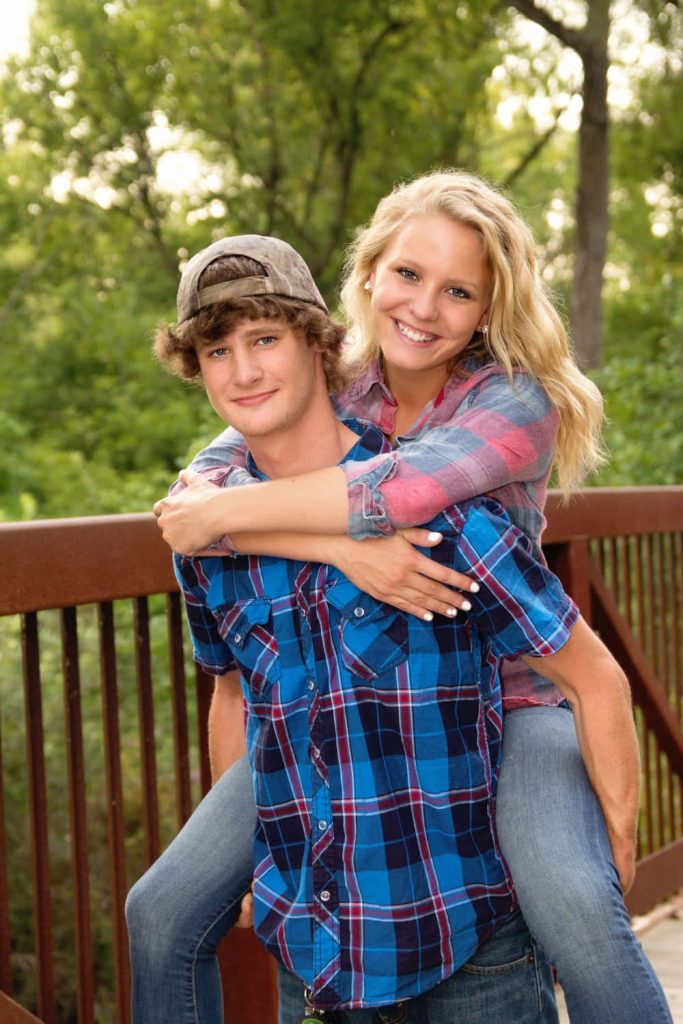 Young couple piggyback ride on park bridge casual plaid outfits lush green romantic wallpaper