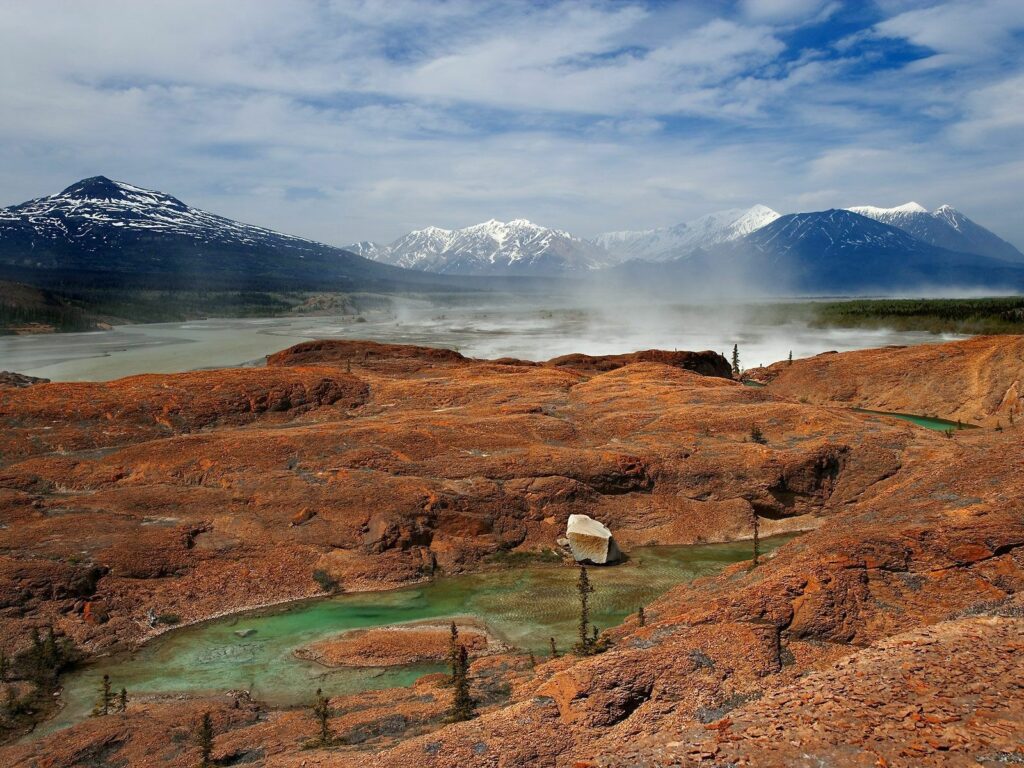 Rugged Canadian landscape with red rocks serene lake steaming geyser snow-capped mountains Wallpaper