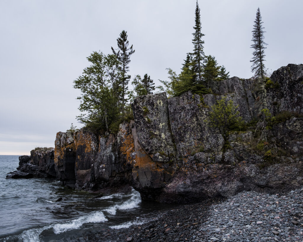 Rugged coastal cliffs with hardy trees by calm gray sea under overcast sky and pebble shore Wallpaper