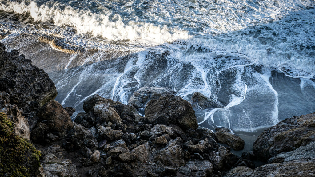 Dynamic coastal wallpaper with rugged stones foamy sea waves and dark wet sand textures