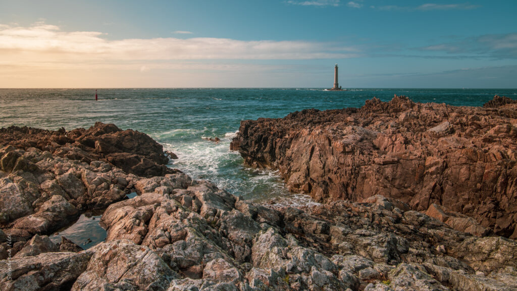 Rugged coastal rocks and blue sea with gentle waves and distant lighthouse under cloudy sky wallpaper