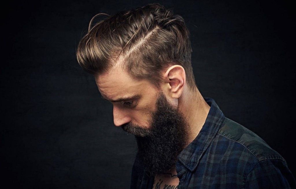 Man with sharp side-part hairstyle and thick beard in plaid shirt against dark background Wallpaper