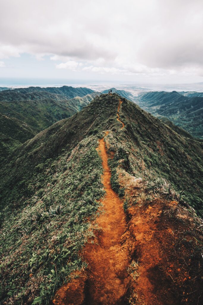 Rugged mountain peak with winding trail and hikers under dramatic sky in lush green landscape Wallpaper