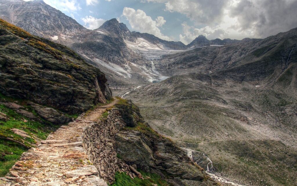Narrow rocky mountain path winding under dramatic sky in vast rugged earthy landscape wallpaper