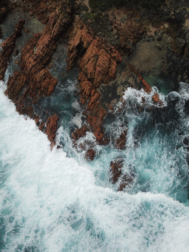 Aerial view of rugged reddish-brown rocks and deep blue sea with foamy waves crashing coastline Wallpaper
