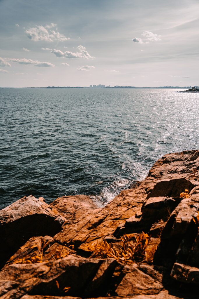 Rugged brown rocks by blue sea with distant city skyline under vast cloud-filled sky background Wallpaper