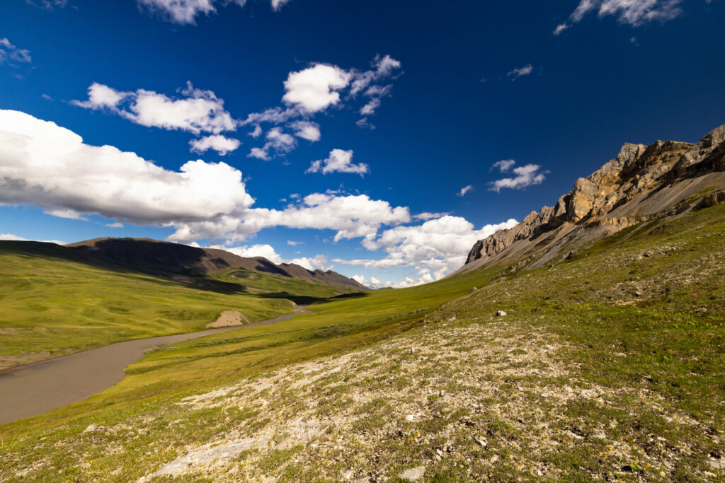Rugged rocks rolling hills lush green field winding river vibrant blue sky fluffy clouds landscape Wallpaper