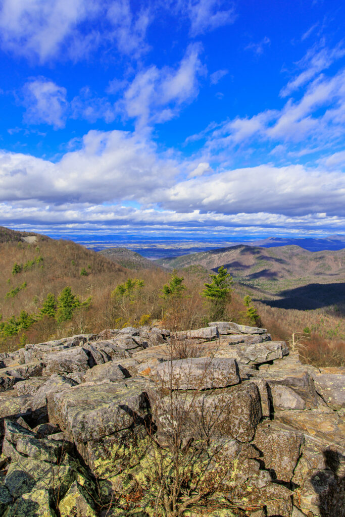 Rugged stone formations with tree-covered mountains and dramatic clouds under blue sky wallpaper