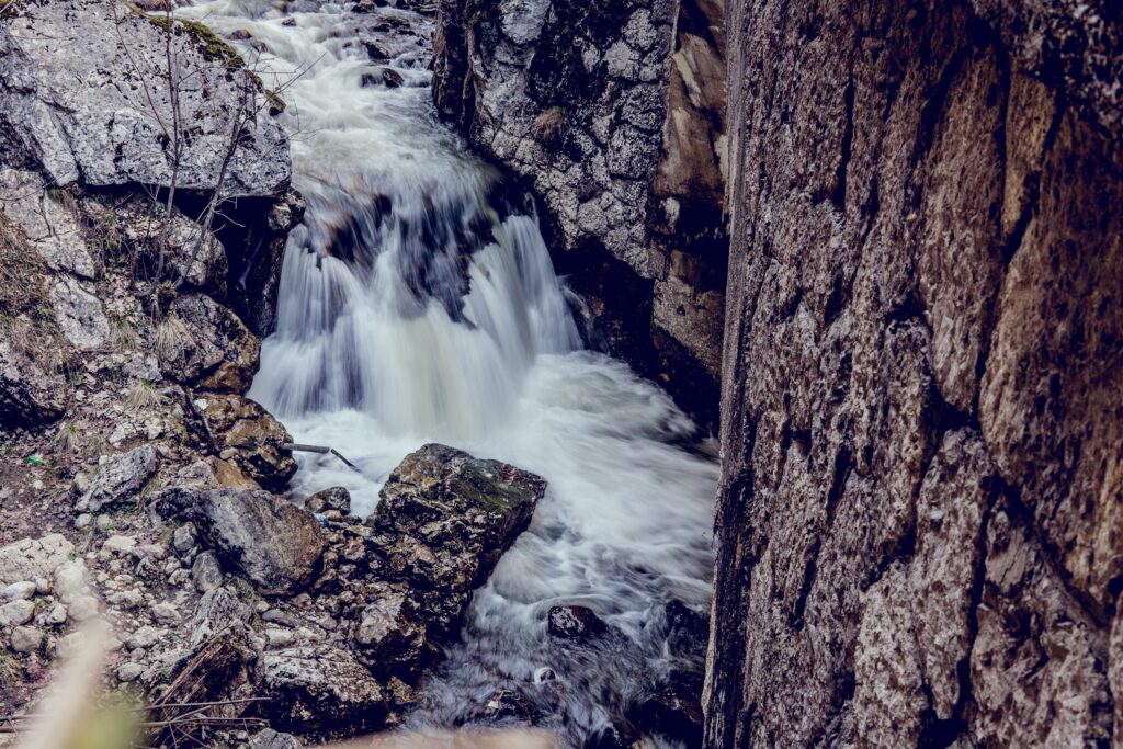 Rushing waterfall over jagged stones flowing into narrow river with textured rocks and earthy tones Wallpaper