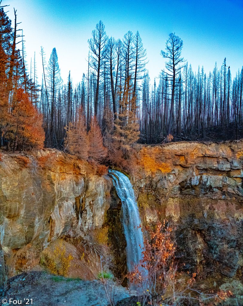 Waterfall cascades from rust-colored cliff with charred forest and bright orange foliage under blue sky Wallpaper