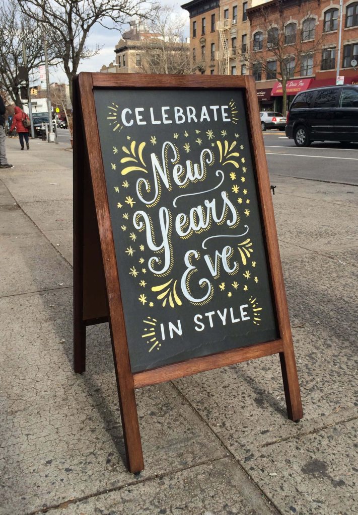 Rustic wooden A-frame blackboard sign on city sidewalk with festive New Years Eve celebration text Wallpaper