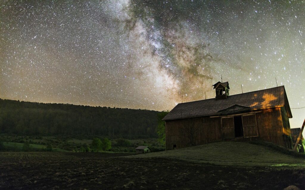 Rustic barn under starry night sky with glowing Milky Way over lush green hills wallpaper