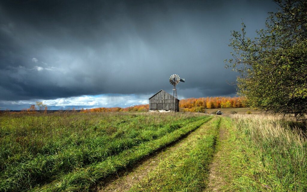 Rustic wooden barn with vintage windmill under dramatic sky and autumn trees in green field Wallpaper