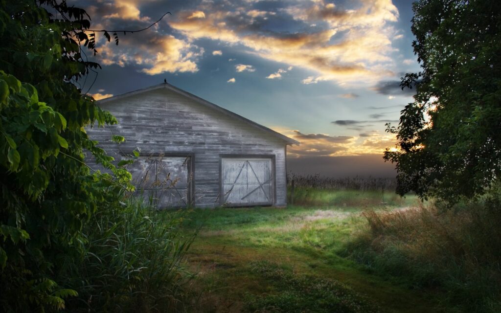 Rustic barn in lush green field under dramatic evening sky with soft sunlight and clouds Wallpaper