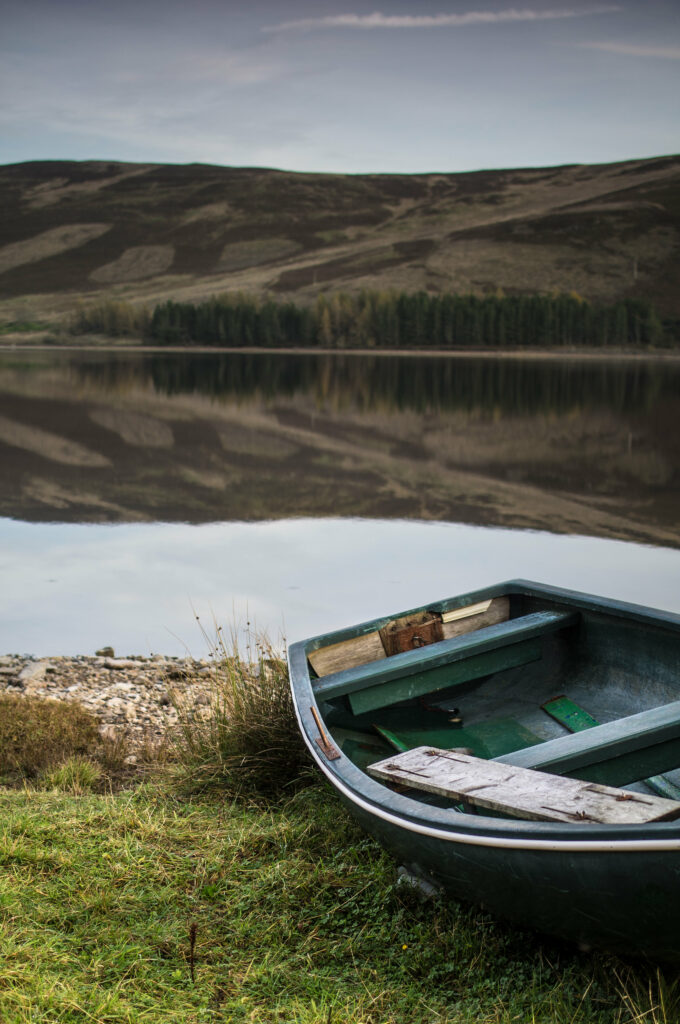 Rustic boat on grassy lake shore reflecting hills and trees under cloudy sky peaceful wallpaper