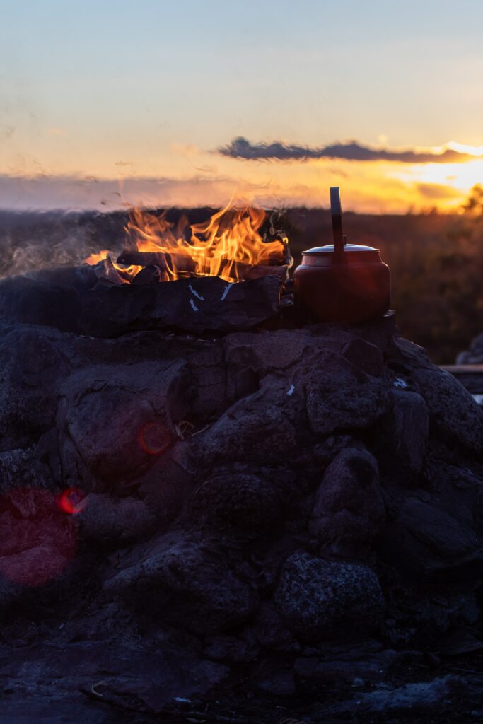 Rustic camping scene with glowing bonfire on stone base and kettle warming at sunset outdoors Wallpaper