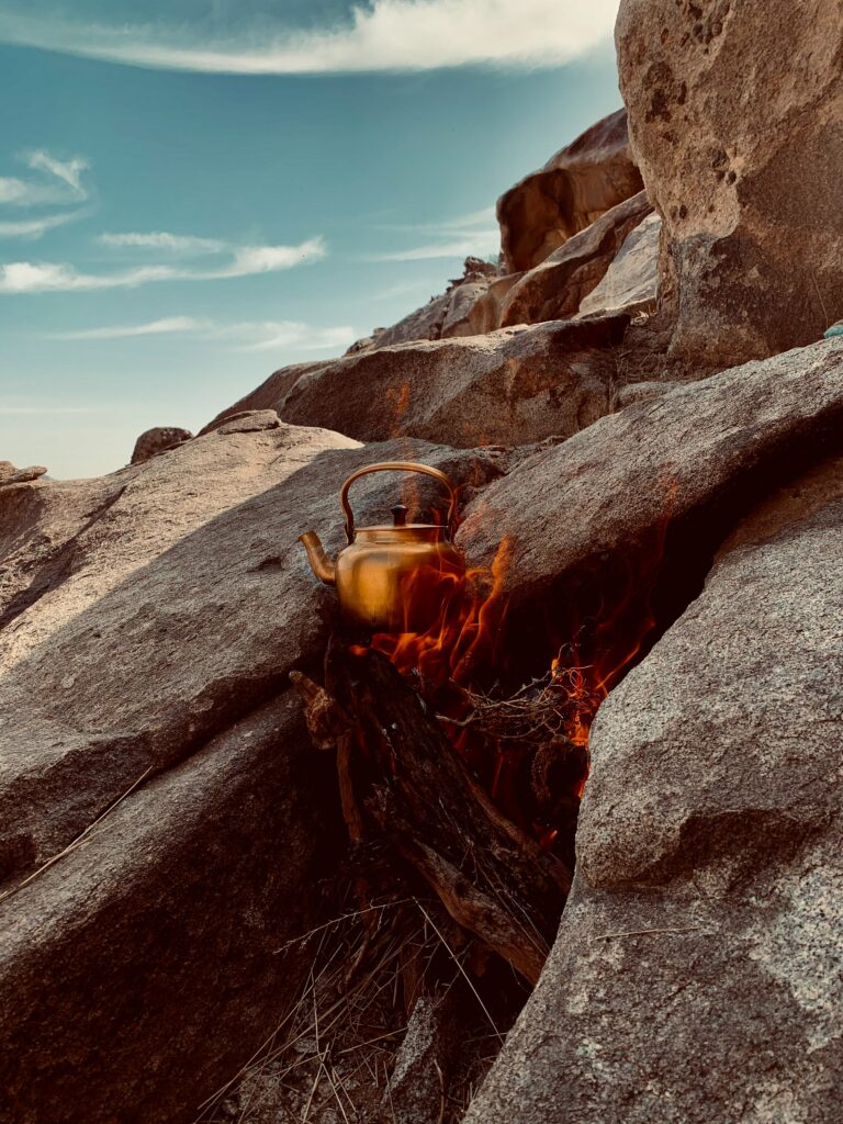 Brass kettle heating over campfire among rugged rocks under clear blue sky outdoors wallpaper