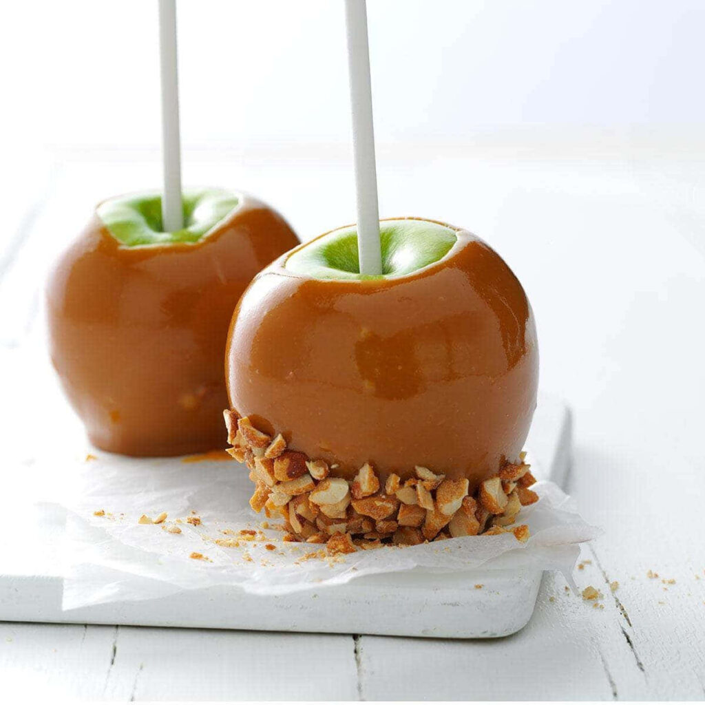 Two glossy caramel apples one coated in crunchy nuts on rustic white wooden table wallpaper background
