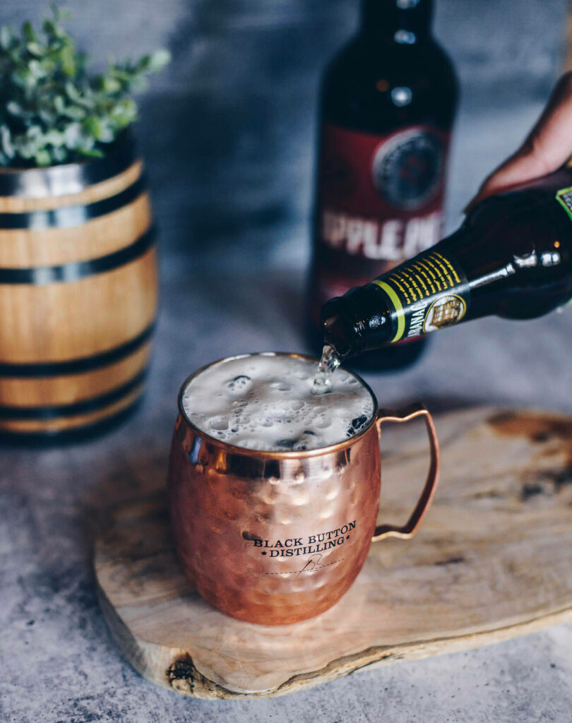 Copper mug labeled Black Button Distilling filled with foamy drink on wooden board with barrel and apple bottle Wallpaper