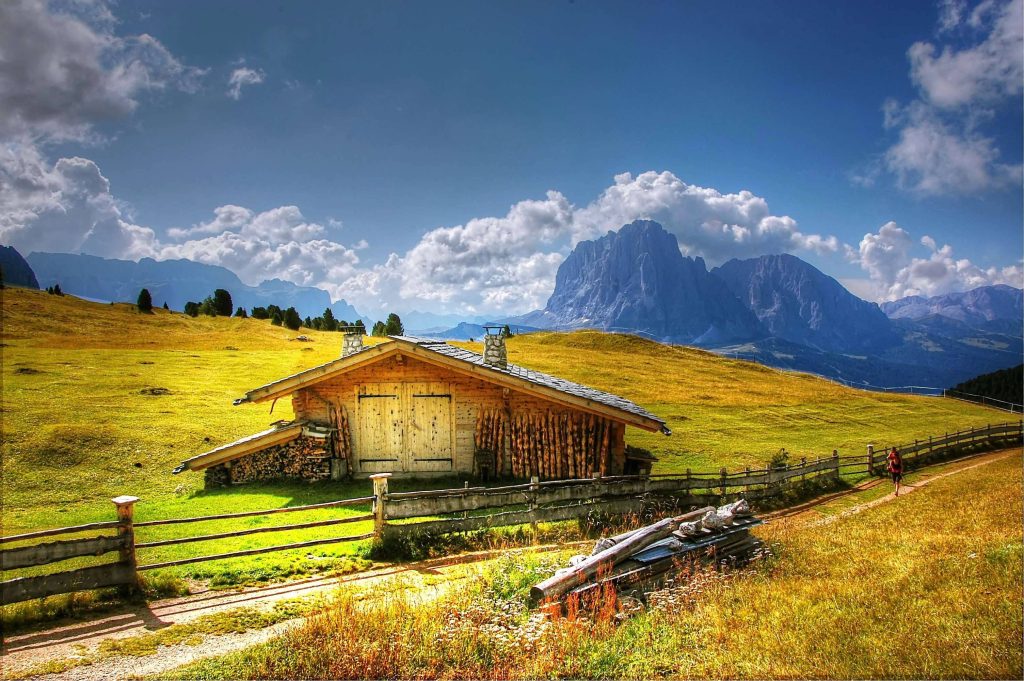 Rustic farm barn and hiker on dirt path with Sassolungo mountains under blue sky autumn landscape Wallpaper