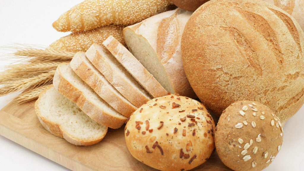 Assorted fresh breads on wooden tray rustic loaves sliced white bread seeded rolls wheat stalks Wallpaper