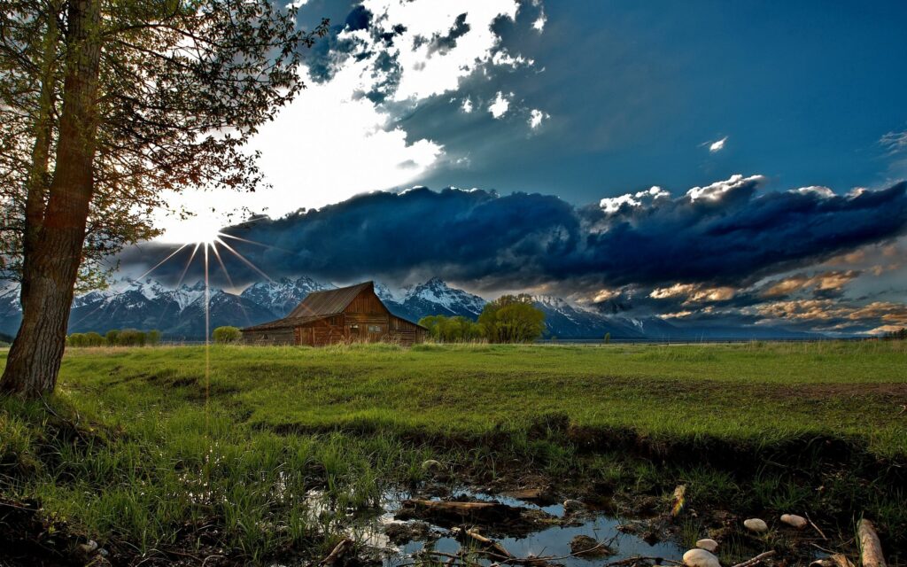 Rustic house in green field with sunbeams dramatic clouds tree mountains and reflective stream wallpaper
