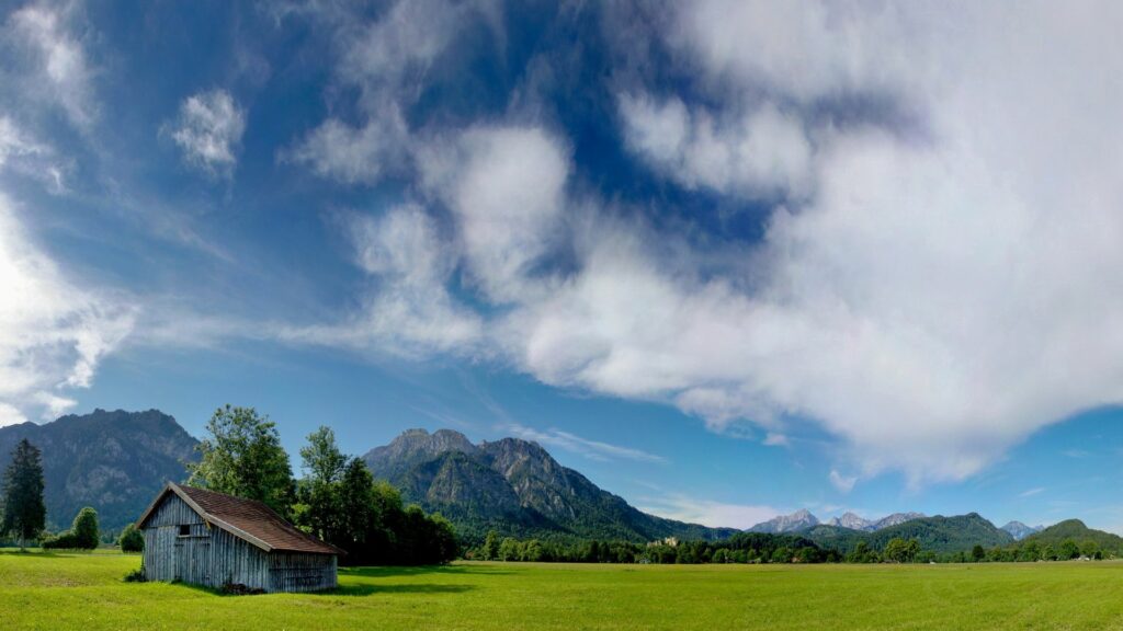 Rustic lodge in green glade beneath cloudy sky with mountains serene peaceful retreat wallpaper