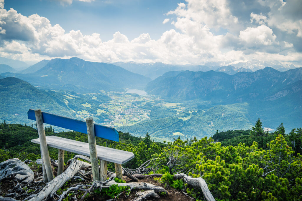 Rustic bench on mountain ridge overlooking lush valleys rolling mountains and cloudy blue sky Wallpaper