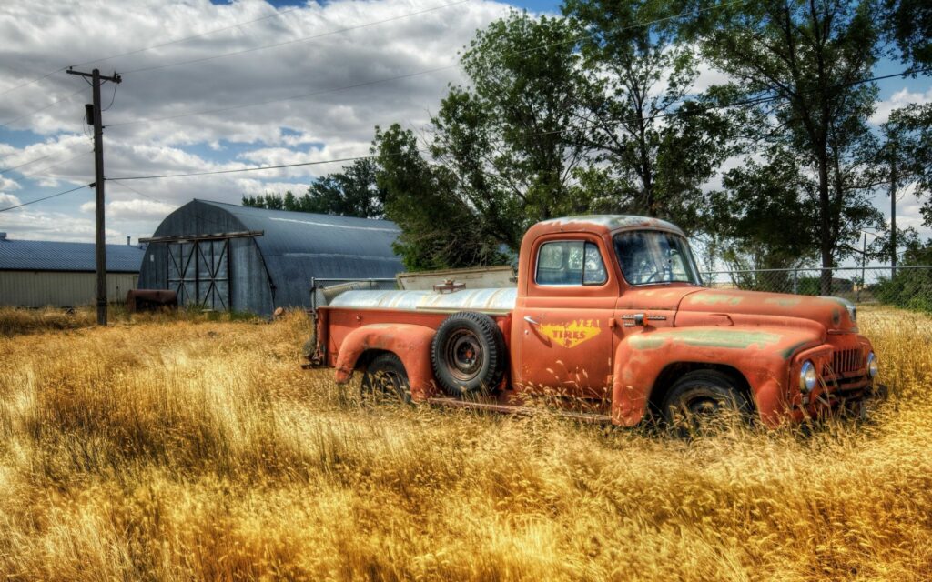 Old red truck with faded paint in golden field near rustic barn and garage under cloudy sky Wallpaper