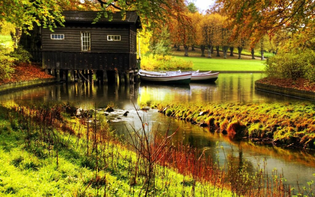 Rustic wooden lodge over calm river with boats lush autumn gardens and sunlit foliage reflections Wallpaper