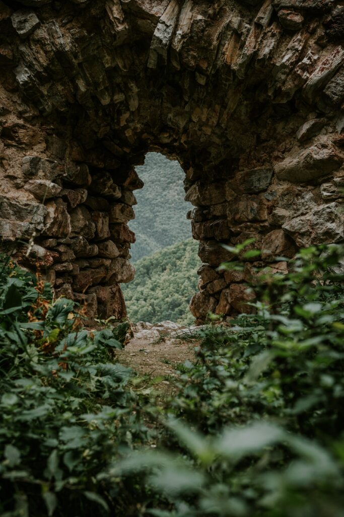 Weathered stone arch amid green grass framing rolling tree-covered hills with rustic textures Wallpaper