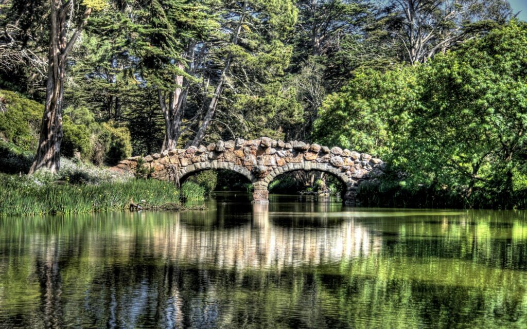 Rustic stone bridge over calm river with vibrant green trees and clear blue sky reflection Wallpaper