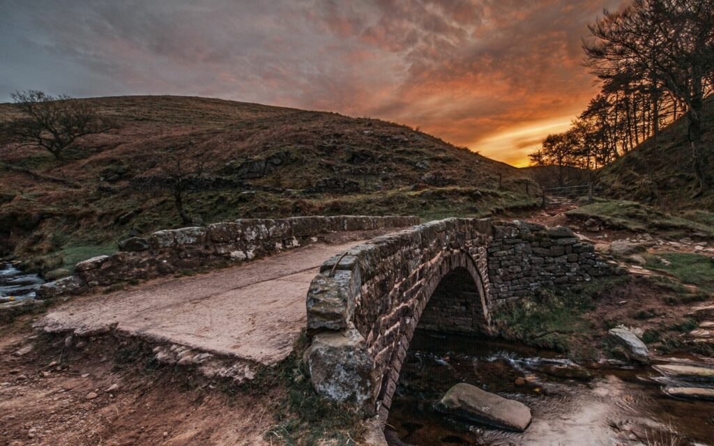 Rustic stone bridge over calm mountain river at sunset with rolling hills and vibrant sky Wallpaper