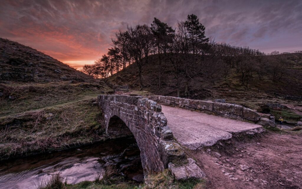 Rustic stone bridge over gentle river with rolling hills and vibrant evening sky wallpaper background