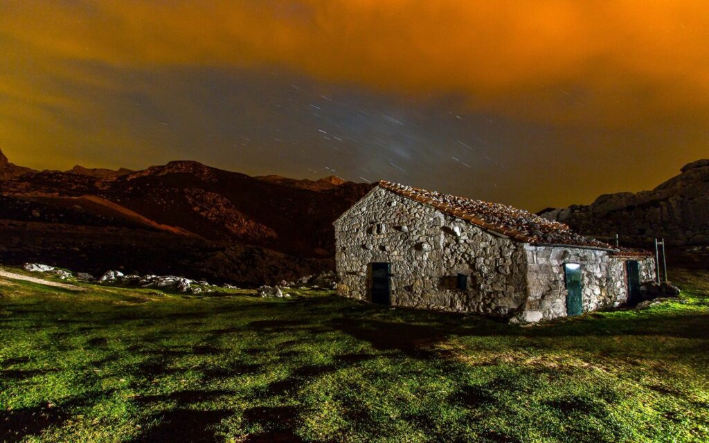 Rustic stone house in grassy field under dramatic orange clouds and starry night sky background Wallpaper