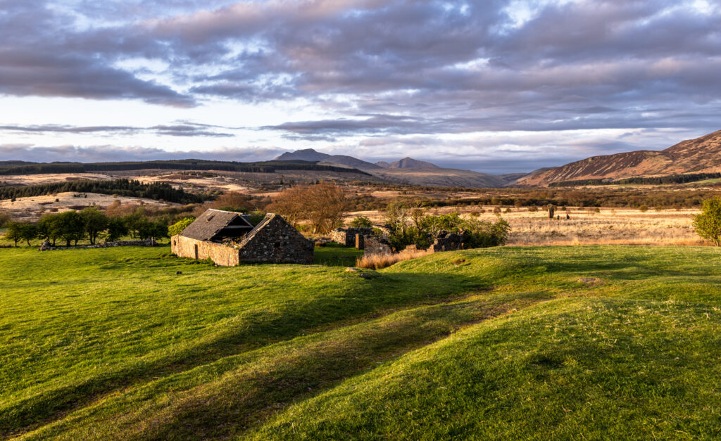 Rustic stone building on green hills under dramatic sky with vibrant grass and distant mountains Wallpaper