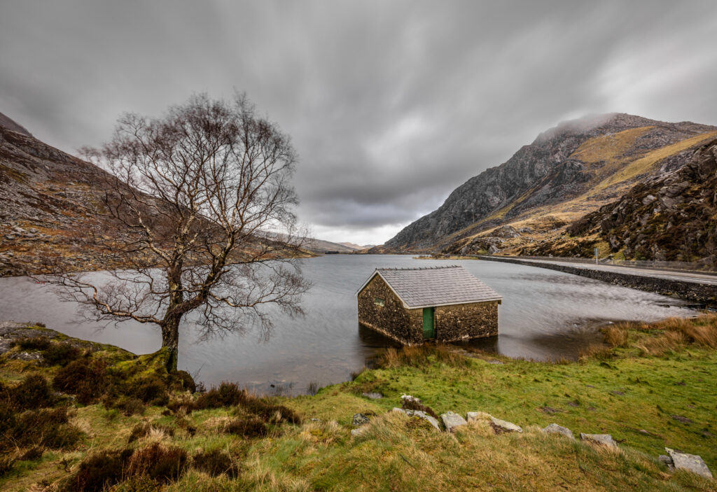 Rustic stone house by calm lake with rugged mountains and solitary tree under cloudy sky wallpaper