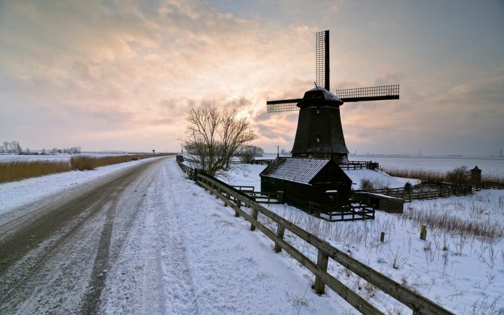 Rustic winter mill by snowy road under cloudy sky with warm sunset in peaceful countryside Wallpaper