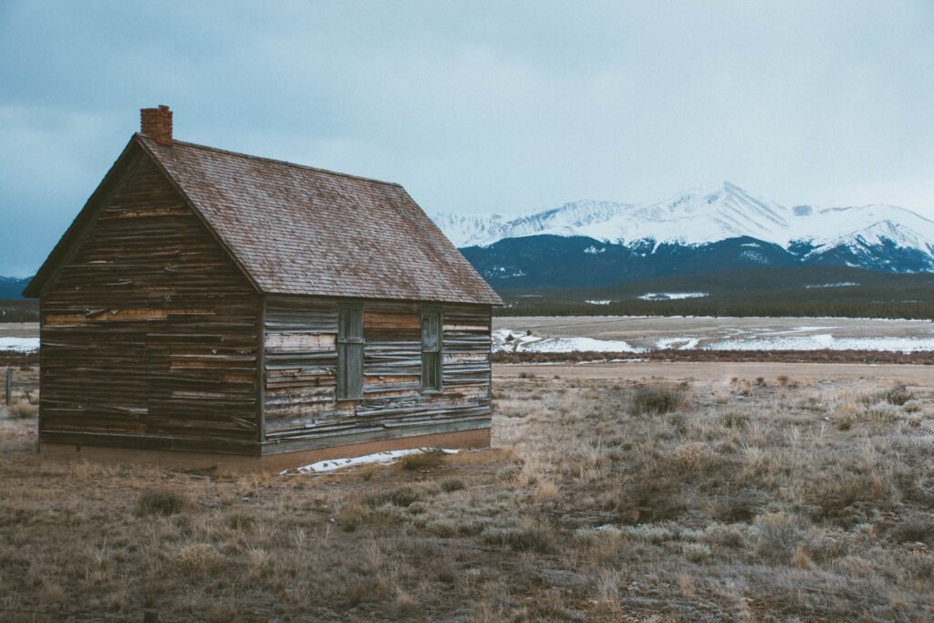 Rustic wooden barn in vast field with snow-capped mountains under cloudy sky rural landscape Wallpaper