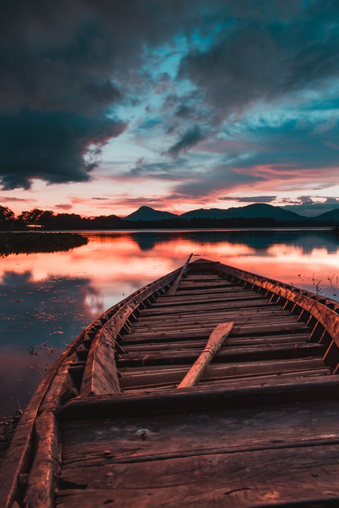 Rustic wooden boat on calm water with vibrant sunset and mountains scenic wallpaper background