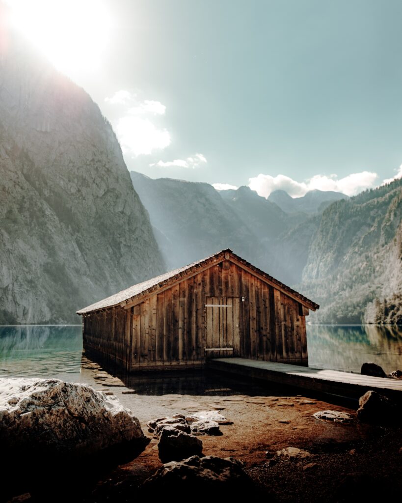 Rustic wooden boathouse by tranquil lake rocky shore dramatic mountain cliffs bright sky wallpaper
