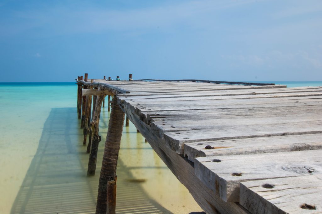 Weathered wooden bridge over clear shallow ocean waters under bright blue sky coastal scene Wallpaper