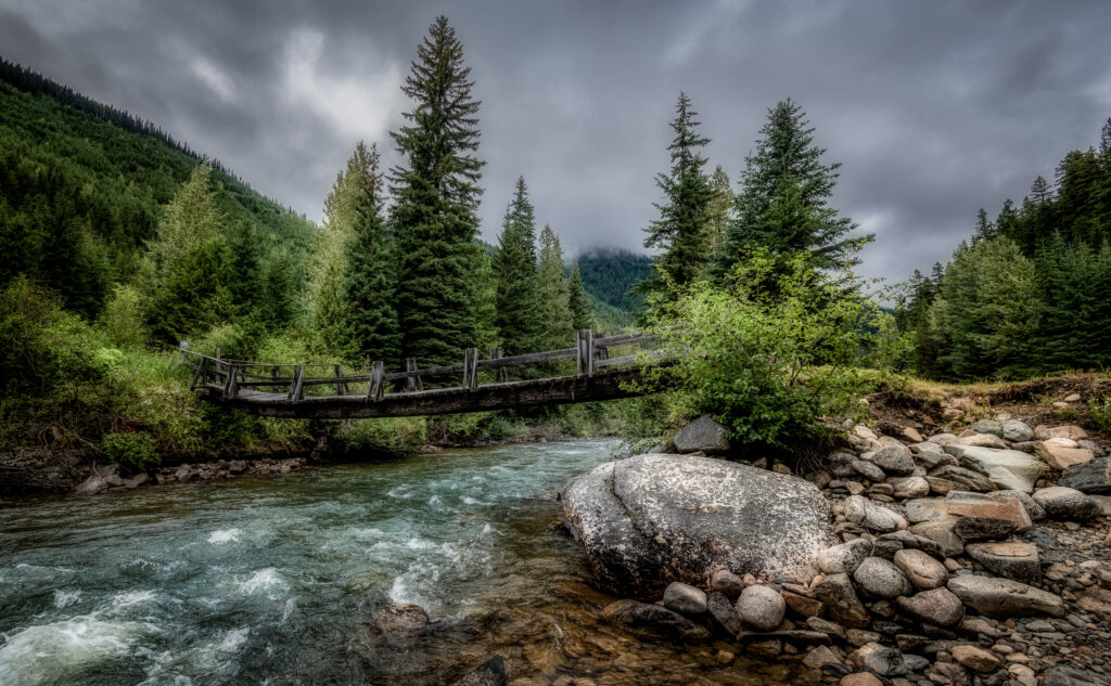 Rustic wooden bridge over rushing river with evergreen trees and cloudy sky wallpaper background