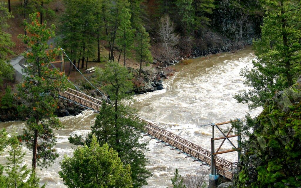 Rustic wooden bridge over rushing river with lush forest and rocky mountains background wallpaper
