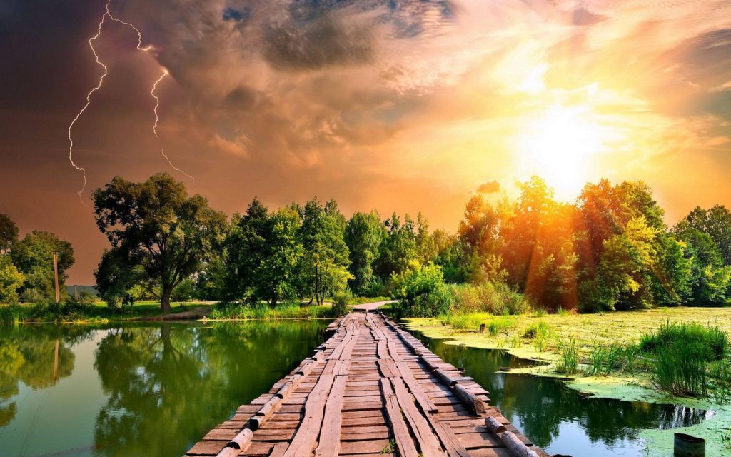 Rustic wooden bridge over serene pond leading to lush forest under stormy sky with lightning Wallpaper